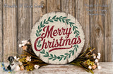 A round wooden sign with the text &quot;Merry Christmas&quot; in red and green lettering, surrounded by a wreath of greenery and flowers, placed on a wooden surface.