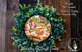 A wreath made of green leaves and flowers, with a teacup and a butterfly resting on it. The wreath is displayed on a wooden surface, and there is a small dog in the bottom right corner.