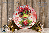 A decorative plate featuring a green frog with red polka dots and a bow, surrounded by flowers and a wooden background.