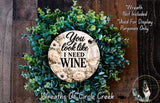 a wreath made of green leaves and flowers, with a circular sign in the center that says "You look like I need wine".