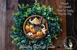 a Halloween-themed wreath made of green foliage and decorated with a pumpkin, ghost, and other spooky elements. The wreath is displayed on a wooden surface, and there is a small dog in the bottom right corner of the image.