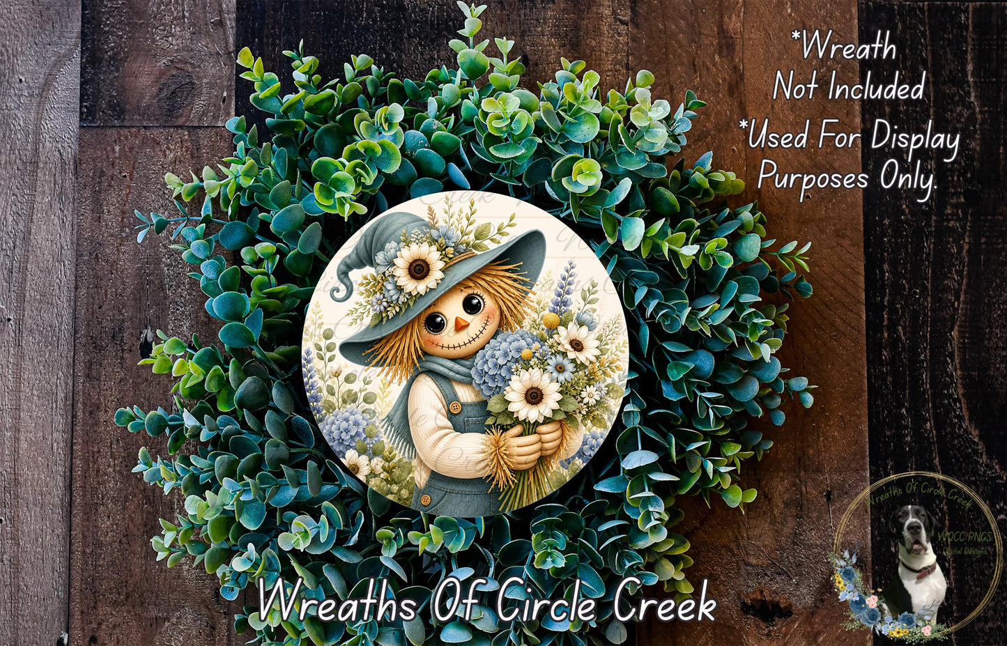a wreath with a scarecrow figure holding flowers, surrounded by a lush green wreath. The wreath is displayed on a wooden surface, and there is a small dog in the bottom right corner of the image.