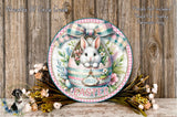 A decorative Easter wreath with a bunny sitting in an Easter egg, surrounded by flowers and greenery. The wreath has a pink and green plaid border and the word "EASTER" written on it.