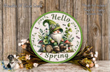 A round sign with a gnome wearing a green hat and surrounded by flowers, mushrooms, and a "Hello Spring" message. The sign is placed on a wooden surface with a floral arrangement nearby.