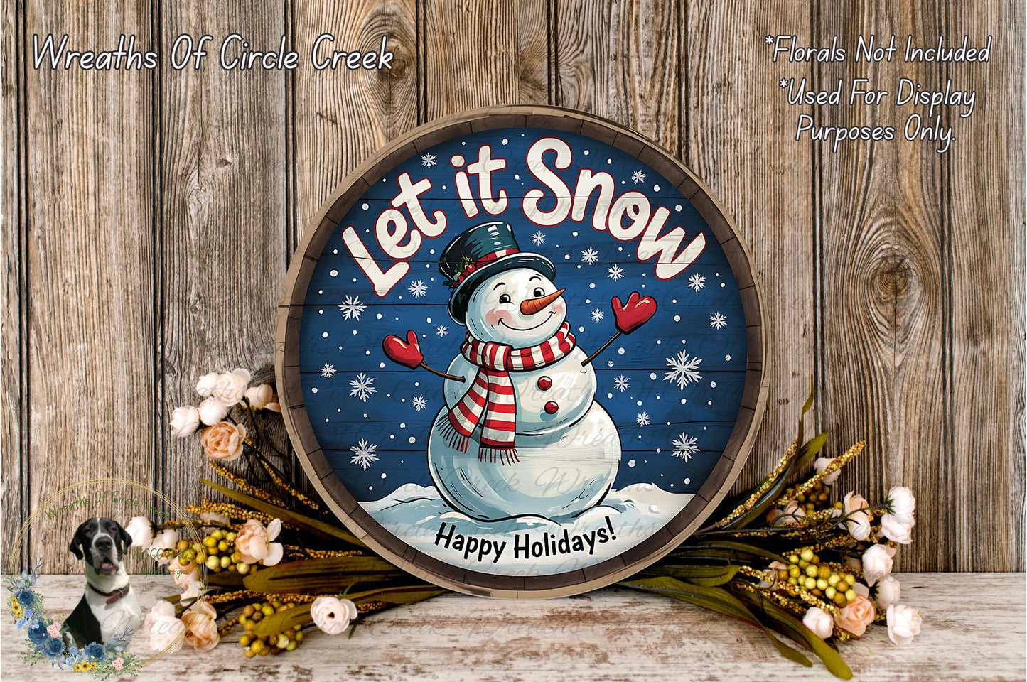 A round wooden sign with a snowman and the text "Let it Snow" is displayed on a wooden surface, surrounded by flowers and greenery.