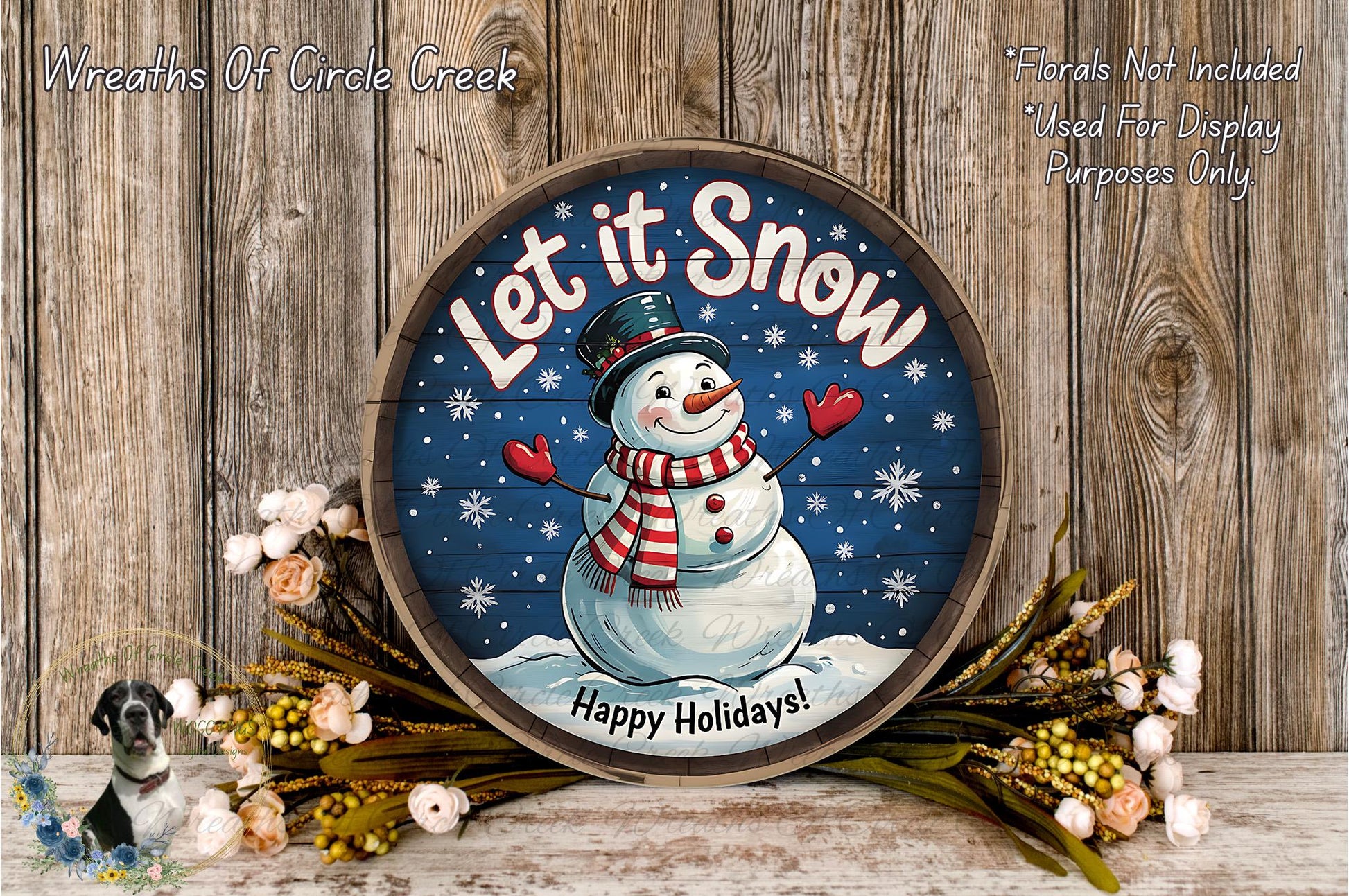 A round wooden sign with a snowman and the text "Let it Snow" is displayed on a wooden surface, surrounded by flowers and greenery.