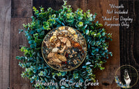 a wreath with a central image of a woman holding a child, surrounded by a lush green plant. The wreath is displayed on a wooden surface, and there is a small dog in the bottom right corner of the image.
