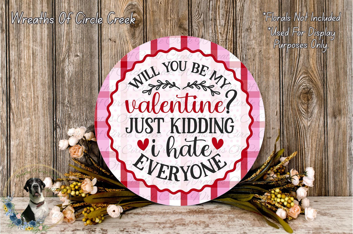 a round sign with a humorous Valentine's Day message, surrounded by a floral arrangement on a wooden surface.