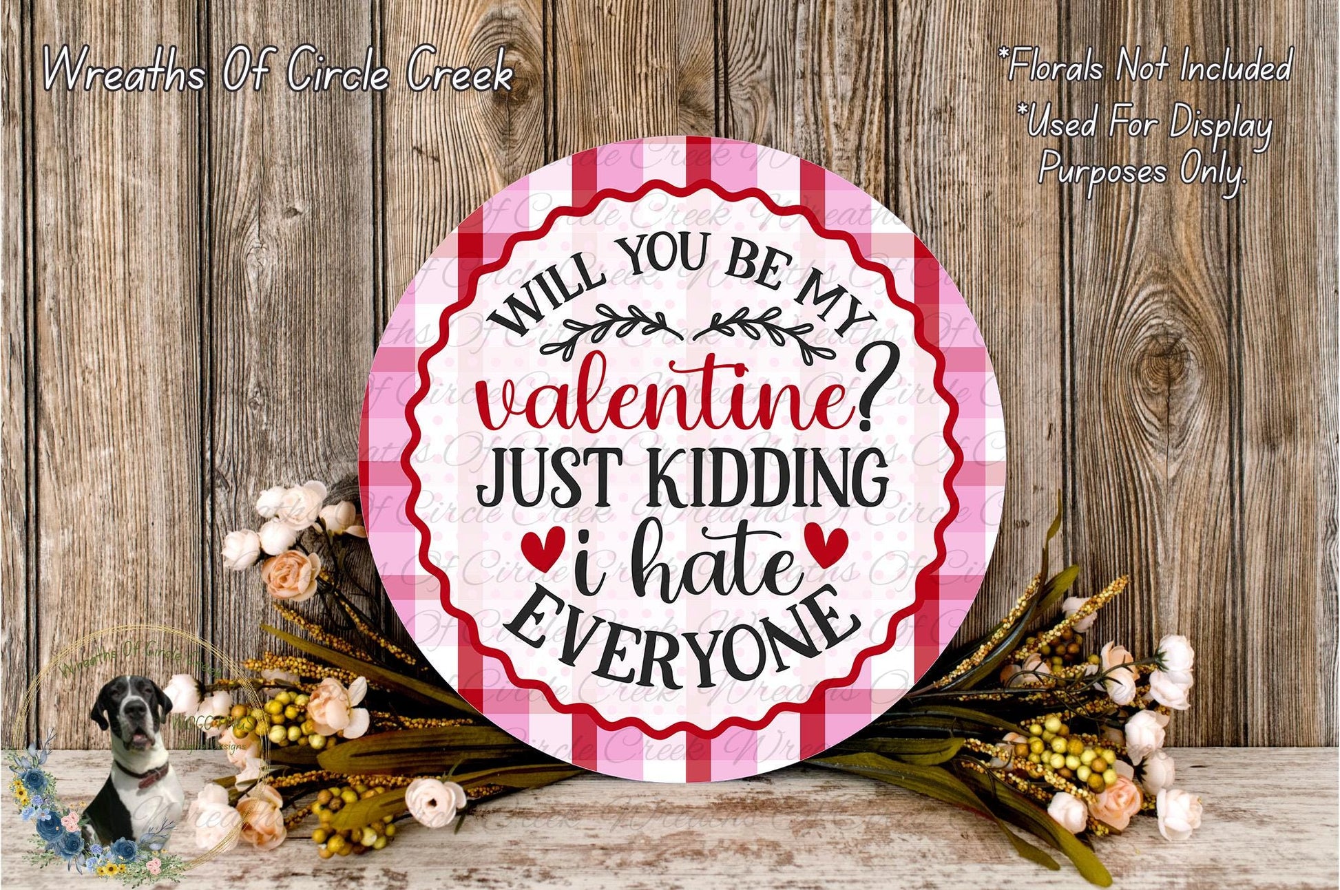 a round sign with a humorous Valentine's Day message, surrounded by a floral arrangement on a wooden surface.