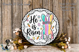 a decorative Easter-themed sign with the text "He is risen" and a colorful illustration of a bunny. The sign is surrounded by a floral arrangement and placed on a wooden surface.