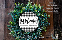 a wreath made of green leaves and a white sign with black text that says "Welcome Just Kidding Please Leave." The wreath is displayed on a wooden surface, and there is a small dog in the bottom right corner of the image.