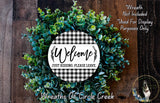 a wreath made of green leaves and a white sign with black text that says "Welcome Just Kidding Please Leave." The wreath is displayed on a wooden surface, and there is a small dog in the bottom right corner of the image.