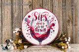a decorative plate with a wine glass and the text "Sips about to go down" written on it, surrounded by flowers and a wooden background.