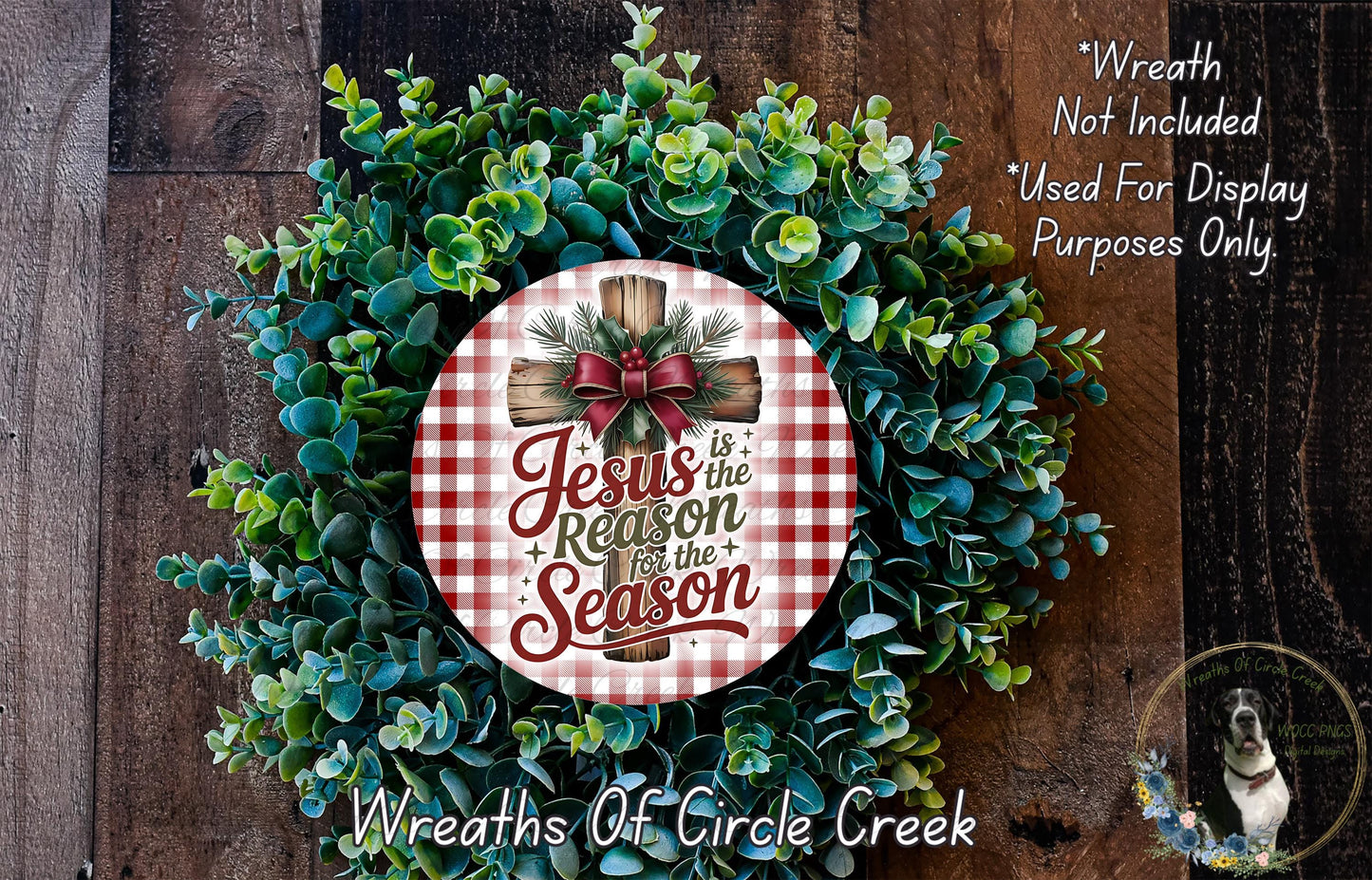 a Christmas wreath with a red and white checkered sign that reads "Jesus is the reason for the season". The wreath is made of green leaves and has a wooden cross at the center. The sign is placed on the wreath, and there is a small dog in the bottom right corner of the image.