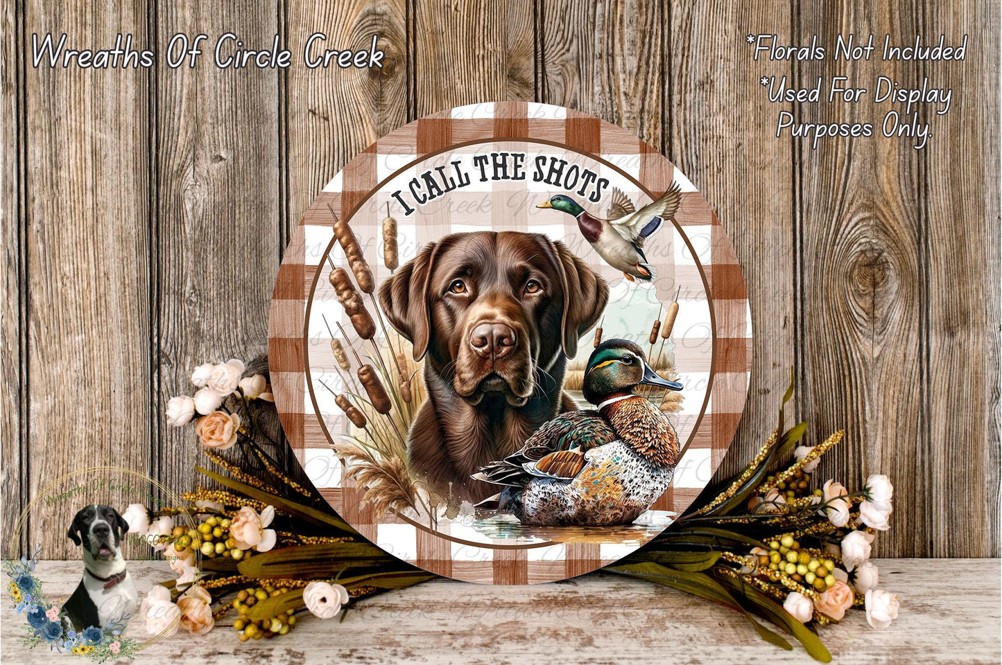A wooden plaque featuring a brown dog and ducks, with the text "I CALL THE SHOTS" and a bird in flight. The plaque is surrounded by flowers and placed on a wooden surface.
