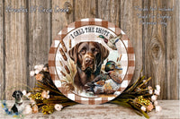 A wooden plaque featuring a brown dog and ducks, with the text "I CALL THE SHOTS" and a bird in flight. The plaque is surrounded by flowers and placed on a wooden surface.