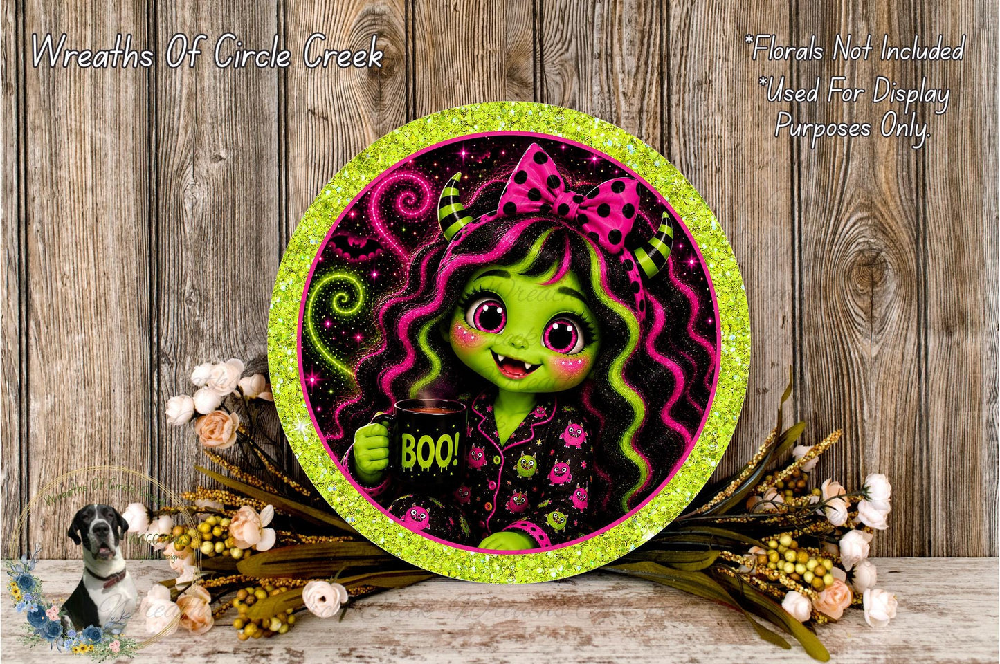 A round, glittery plate featuring a cartoon image of a green-skinned, pink-haired girl with a bow, holding a "Boo!" sign. The plate is surrounded by a floral arrangement and placed on a wooden surface.
