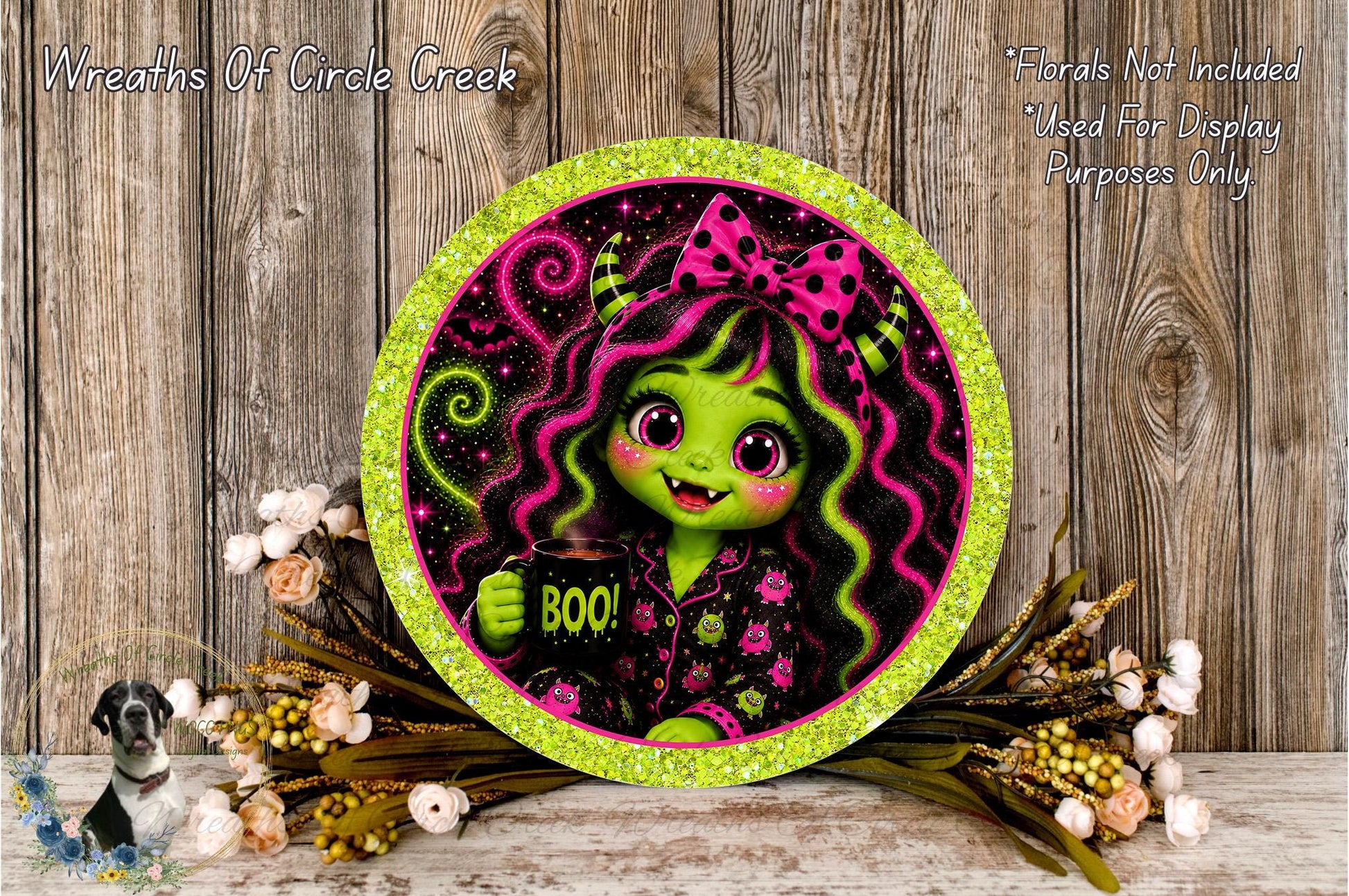 A round, glittery plate featuring a cartoon image of a green-skinned, pink-haired girl with a bow, holding a "Boo!" sign. The plate is surrounded by a floral arrangement and placed on a wooden surface.