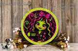 A round, glittery plate featuring a cartoon image of a green-skinned, pink-haired girl with a bow, holding a "Boo!" sign. The plate is surrounded by a floral arrangement and placed on a wooden surface.