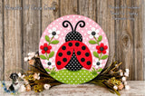 A decorative round sign featuring a red ladybug surrounded by flowers and green leaves, placed on a wooden surface.