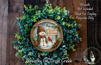 A wreath with a snowman and a sign that says "Merry Christmas" is displayed on a wooden background.