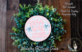 a wreath made of green leaves and a pink circular sign with the text "Blush your heart" on it, placed on a wooden surface.