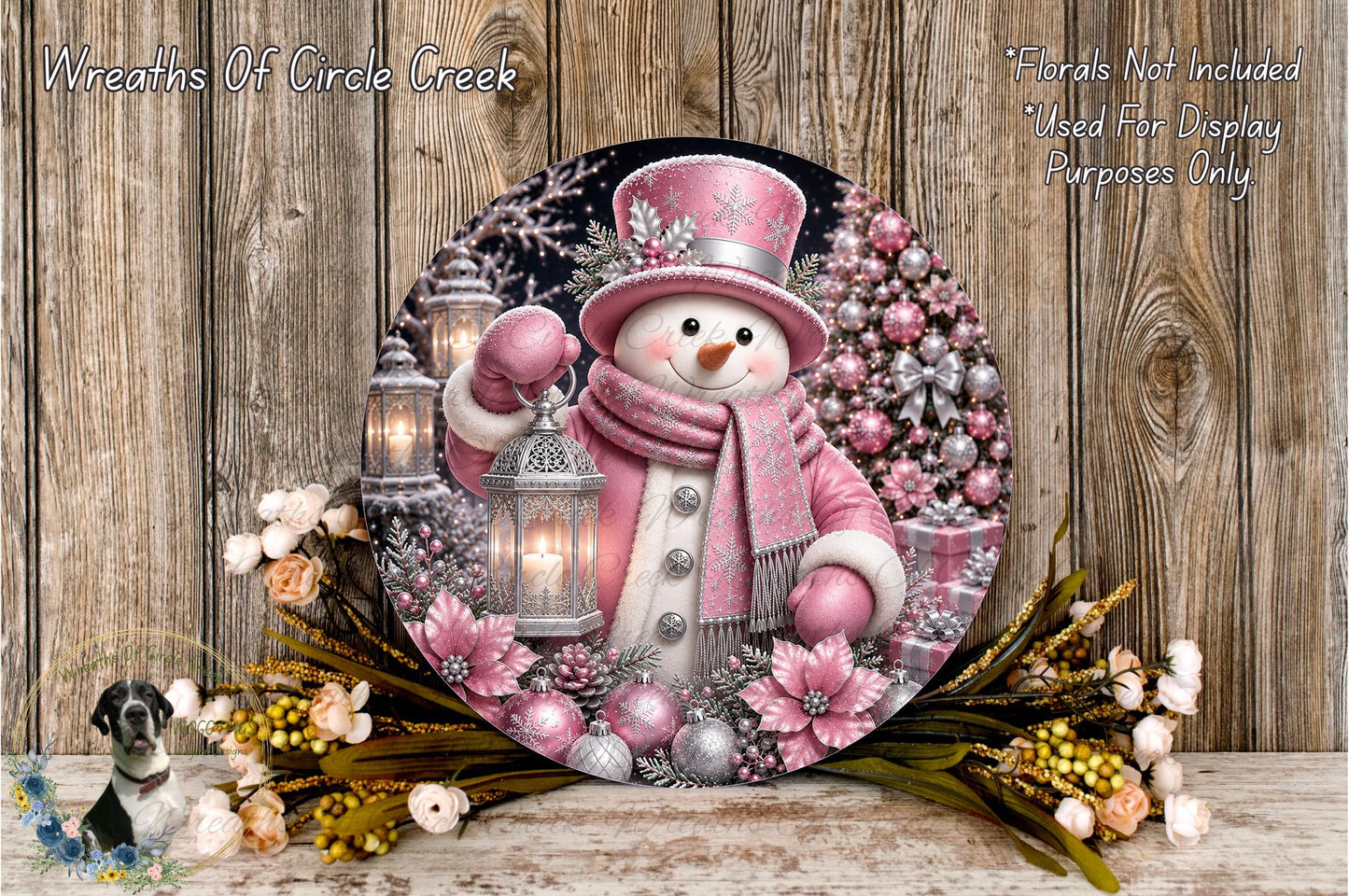 A snowman wearing a pink outfit and top hat, holding a lantern, surrounded by a wreath of pink flowers and a Christmas tree, with a wooden background.