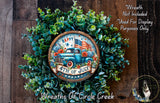 A wreath made of green leaves and flowers, with a circular sign featuring a blue truck and an American flag, hanging on a wooden wall.