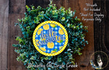 A wreath made of green leaves and topped with a yellow and blue circular sign that says "Hello summer".