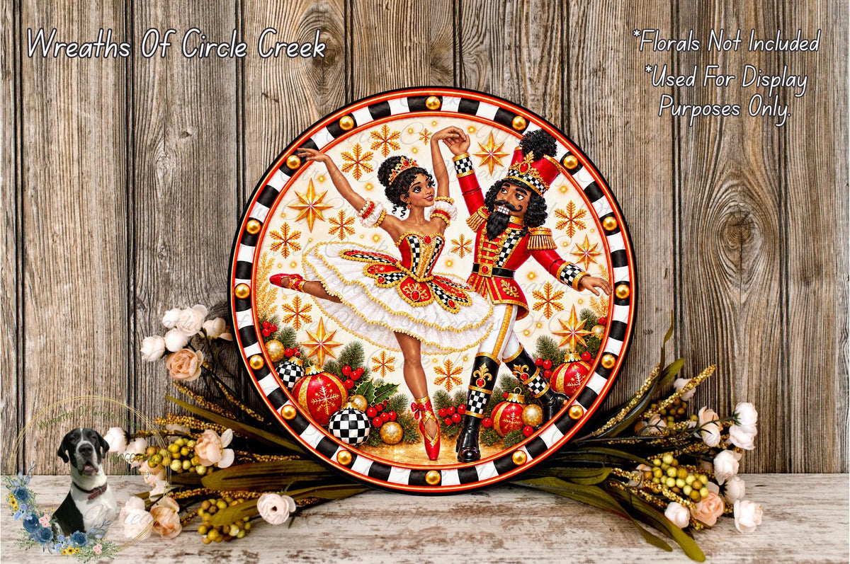 a decorative plate with a painting of two dancers, one in a white tutu and the other in a red and gold costume, surrounded by a wreath of flowers and a wooden background.