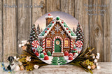 A round wooden plaque depicting a gingerbread house with a snowy landscape, surrounded by flowers and greenery.