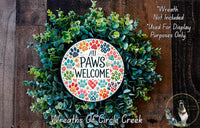 A wreath made of various green plants and colorful paw prints, with a circular sign that says &quot;All Paws Welcome&quot; in the center.
