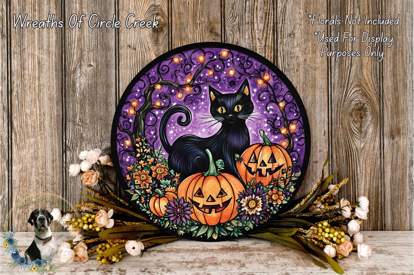 A Halloween-themed wreath featuring a black cat, two jack-o'-lanterns, and a purple background with a tree and flowers. The wreath is displayed on a wooden surface, surrounded by flowers and greenery.