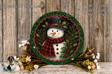 A snowman wearing a red and black plaid scarf and a black top hat, surrounded by a green wreath and flowers, is displayed on a wooden surface.