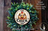 a wreath with a large, whimsical chicken face design at the center, surrounded by green foliage. The wreath is displayed on a wooden surface, and there is a small dog visible in the bottom right corner of the image.