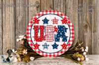 a circular sign with the word "USA" written on it, surrounded by various patriotic symbols such as stars, stripes, and flowers. The sign is placed on a wooden surface, and there are some flowers and greenery nearby.