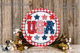 a circular sign with the word "USA" written on it, surrounded by various patriotic symbols such as stars, stripes, and flowers. The sign is placed on a wooden surface, and there are some flowers and greenery nearby.