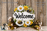 A decorative welcome sign with a floral design, featuring a bee and flowers, placed on a wooden surface.
