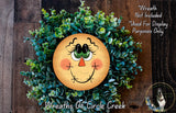 a wreath with a smiling face made of greenery, including small leaves and flowers. The wreath is displayed on a wooden surface, and there is a small dog in the bottom right corner of the image.
