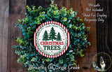 a Christmas wreath made of evergreen branches and a circular sign with the text "Christmas Trees" on it, placed on a wooden surface.
