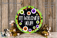a Halloween-themed sign with the text "I'm watching you!!!" displayed on a green circular background. The sign is surrounded by a floral arrangement and placed on a wooden surface.