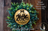 A wreath made of green leaves and a golden circular ornament with the text "Hello 2006" on it, placed on a wooden surface.
