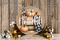 a round wooden sign with the word "Thankful" written on it, surrounded by autumn-themed decorations such as pumpkins, flowers, and a plaid cloth.