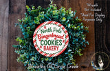 A wreath made of green leaves and a red and white sign that says "North Pole Gingerbread Cookies Bakery" is displayed on a wooden surface.