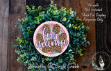 a wreath made of various green plants and a pink circular sign with the text "fur mama" written on it. The wreath is displayed on a wooden surface, and there is a small dog visible in the bottom right corner of the image.