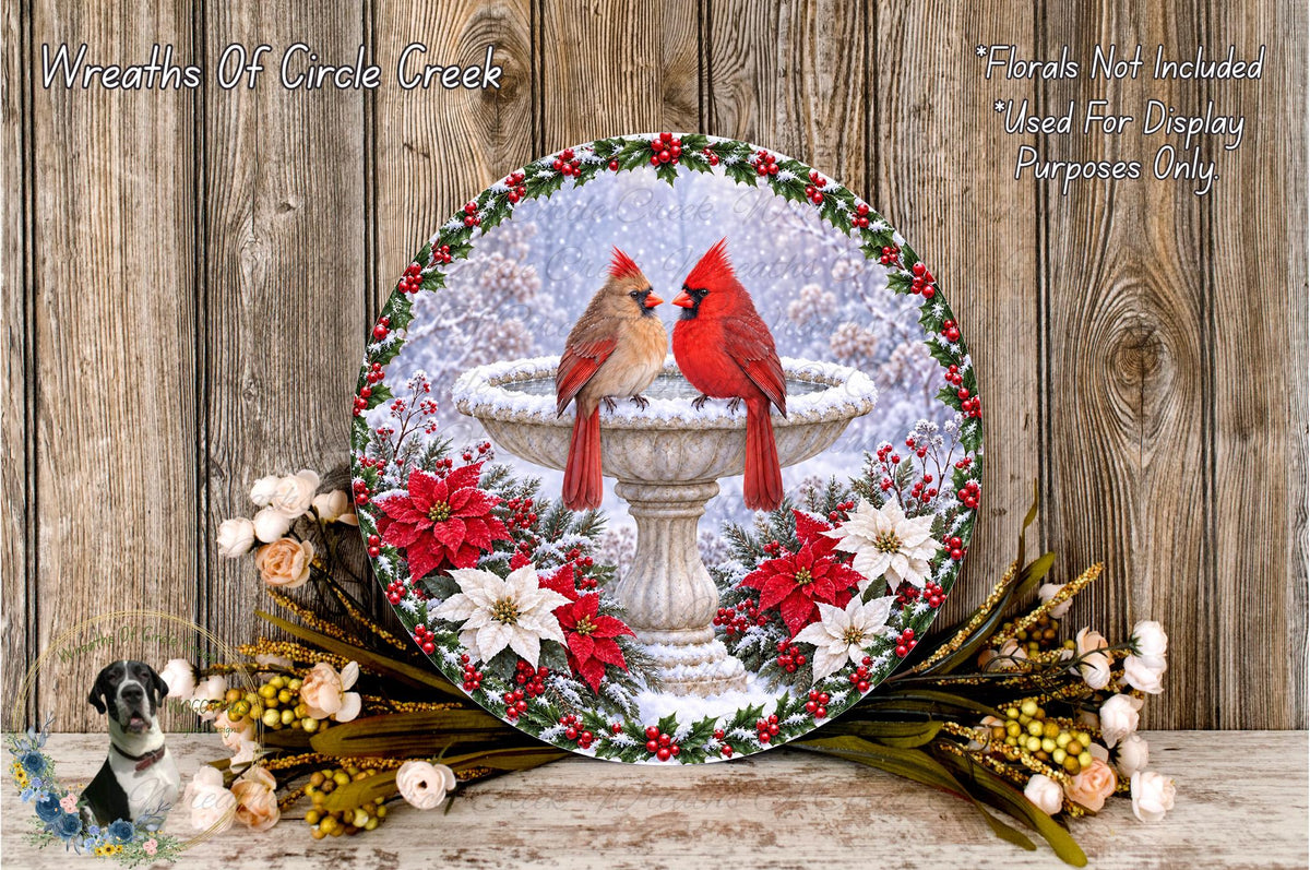 A decorative wreath featuring two red cardinals perched on a fountain, surrounded by poinsettia flowers and greenery, set against a snowy background.