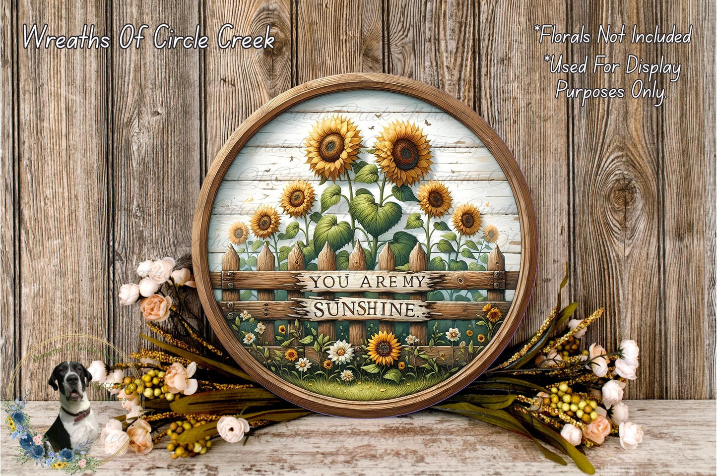 A round wooden frame with a painting of a sunflower garden and the text "You are my sunshine" displayed on it, surrounded by a wreath of flowers and a wooden fence.