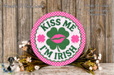 a circular sign with the text "KISS ME I'M IRISH" and a shamrock graphic, surrounded by a floral arrangement on a wooden surface.