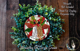 A wreath made of various green plants and a red ribbon with the word "Believe" written on it, placed on a wooden surface.
