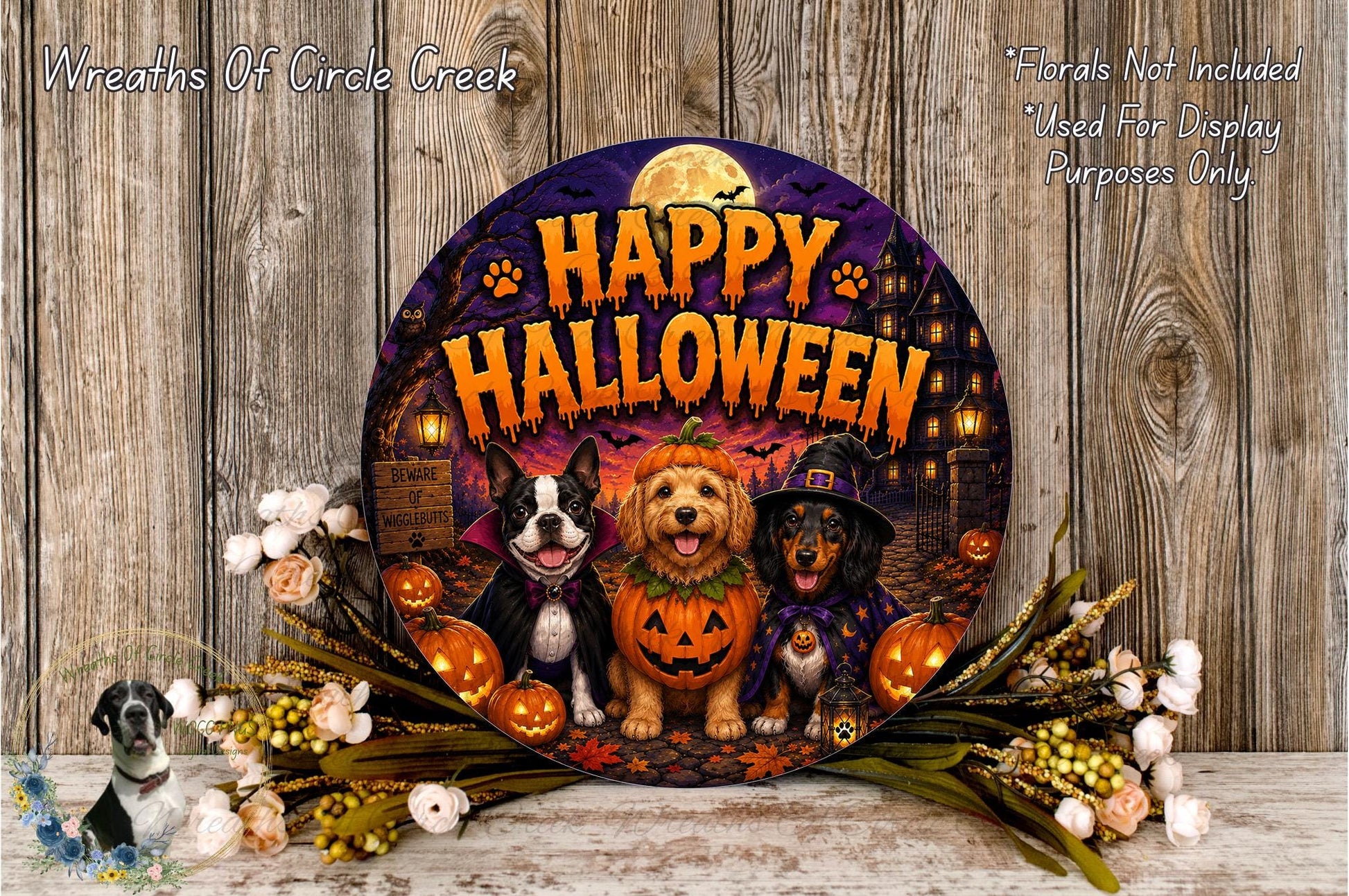A Halloween-themed wreath featuring three dogs dressed in costumes, with a jack-o-lantern and other Halloween-related elements. The wreath is surrounded by flowers and placed on a wooden surface.
